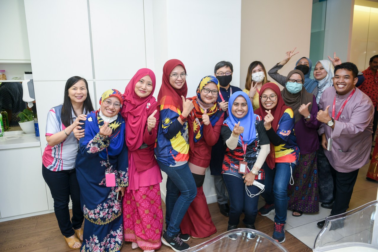Colleagues in patriotic Malaysia-themed outfits celebrating Merdeka with joy during an office lunch catering event.